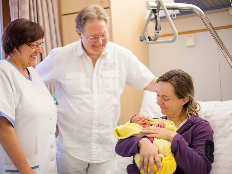 Zwei Ärzte reden mit einer Frau, die im Patientenbett sitzt und ihr neugeborenes Baby im Arm hält